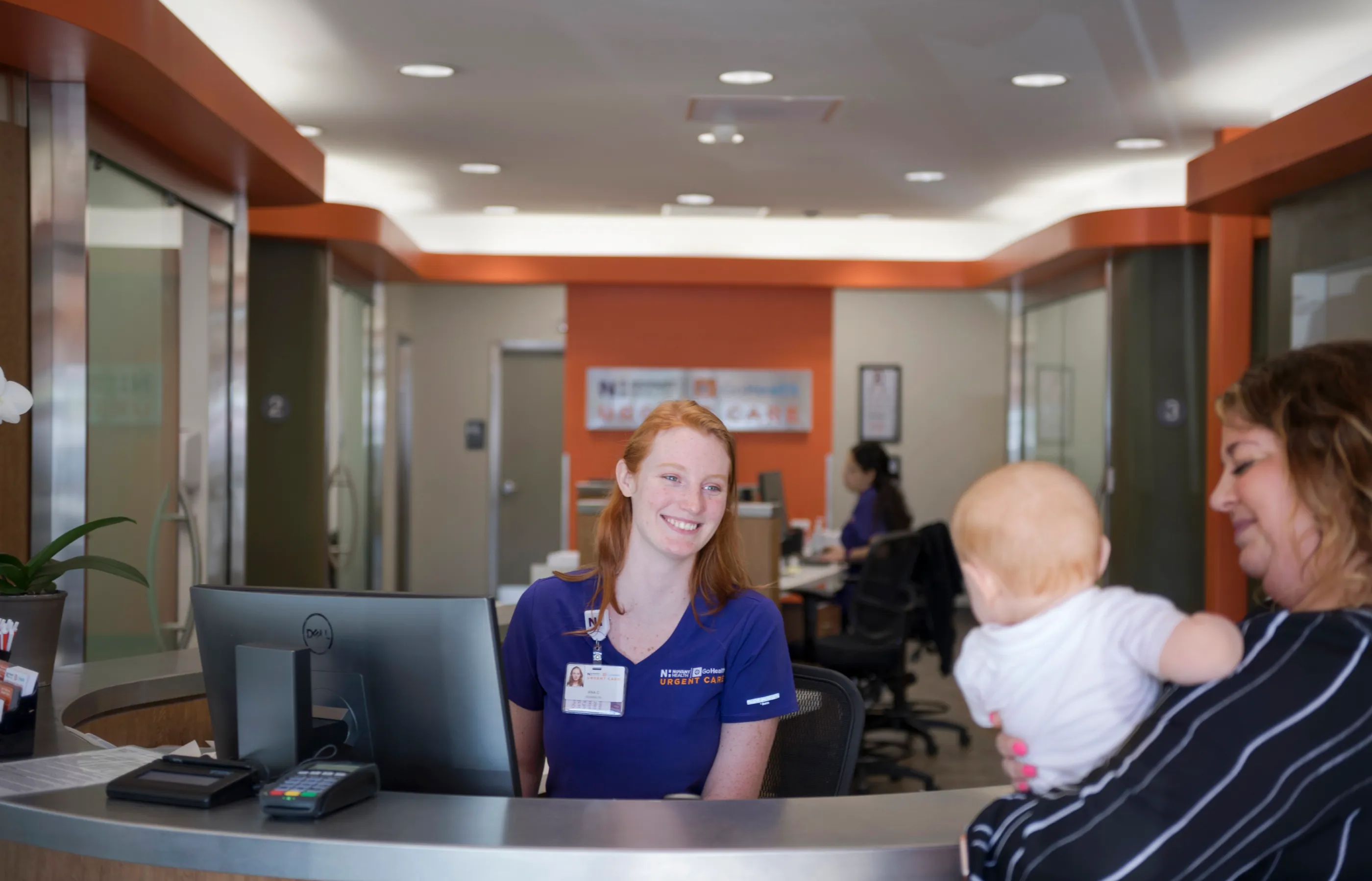 A Novant Health team member is greeting a mother and her baby at the clinic's front desk. 