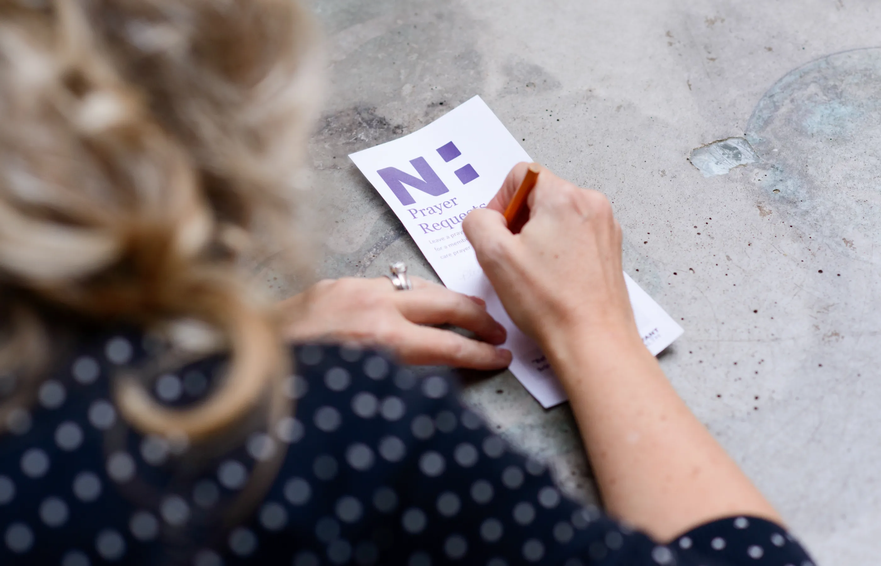 Woman is sitting with a pen and a Novant Health prayer request form. She's writing in her prayer request for a Novant Health patient. 