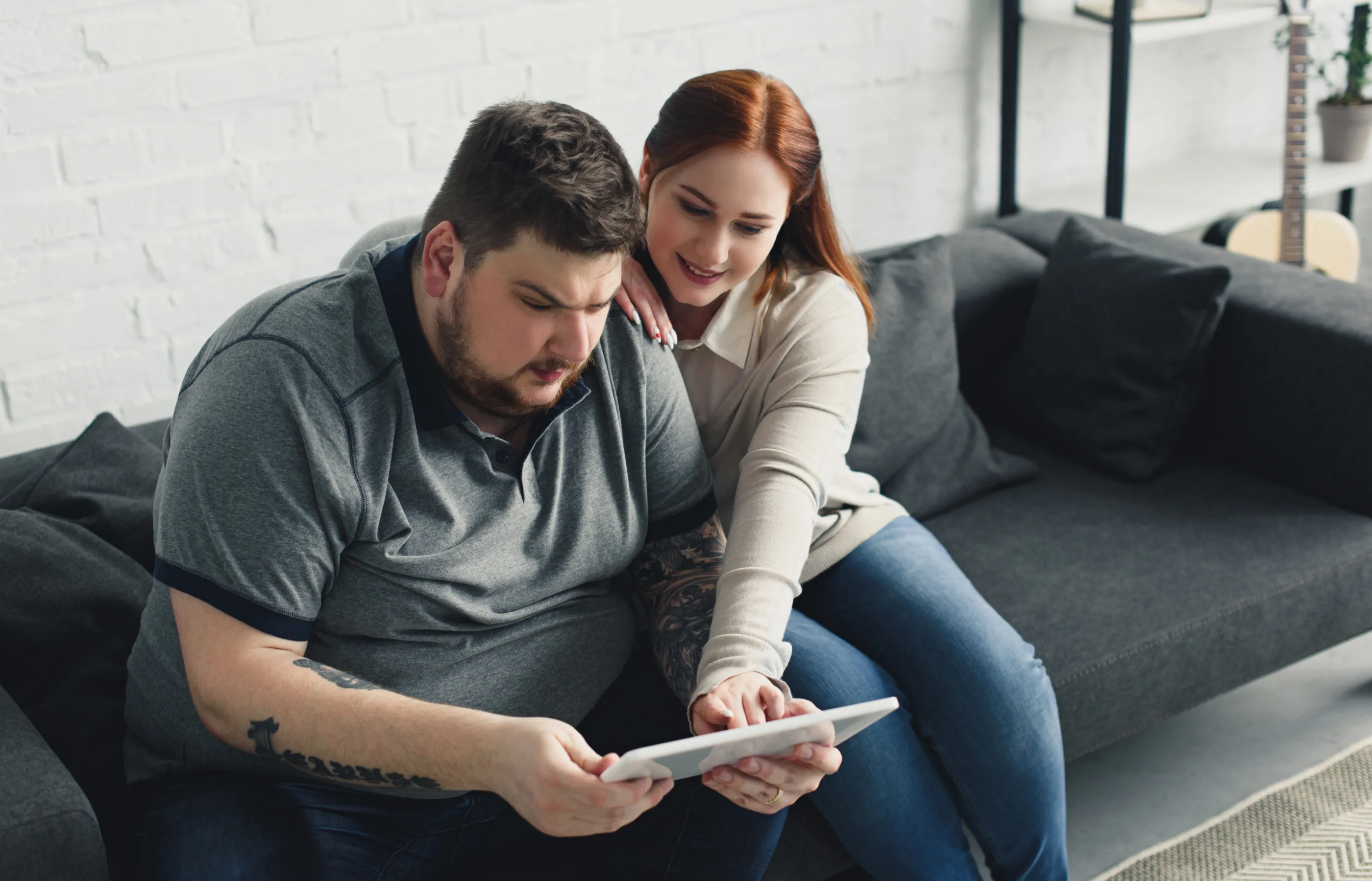 A man is sitting on the couch with his partner. He is holding a tablet and reviewing information, as his partner leans over to point at something on the screen. 