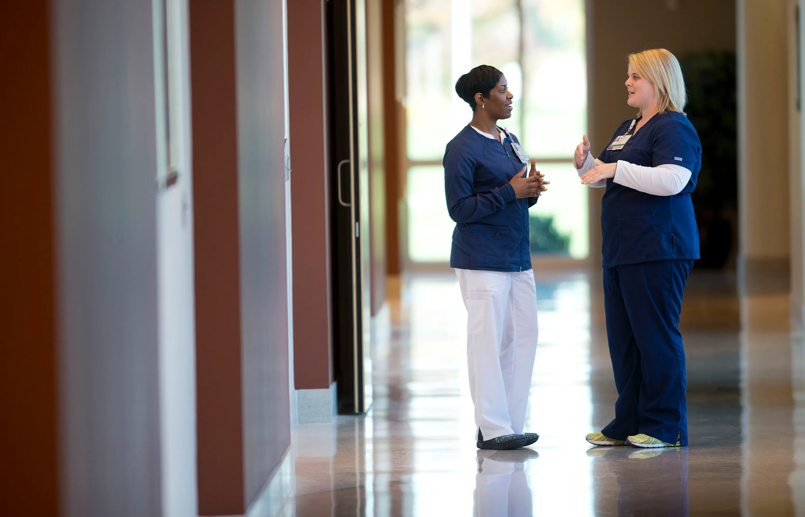 Two Novant Health registered nurses are standing in the hall of a medical center talking. 